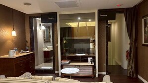 A hotel room with a sofa, a small round table, a minibar, a TV, and a glass partition separating the living area from the bathroom. - Langkei Hotel (Taoyuan City)