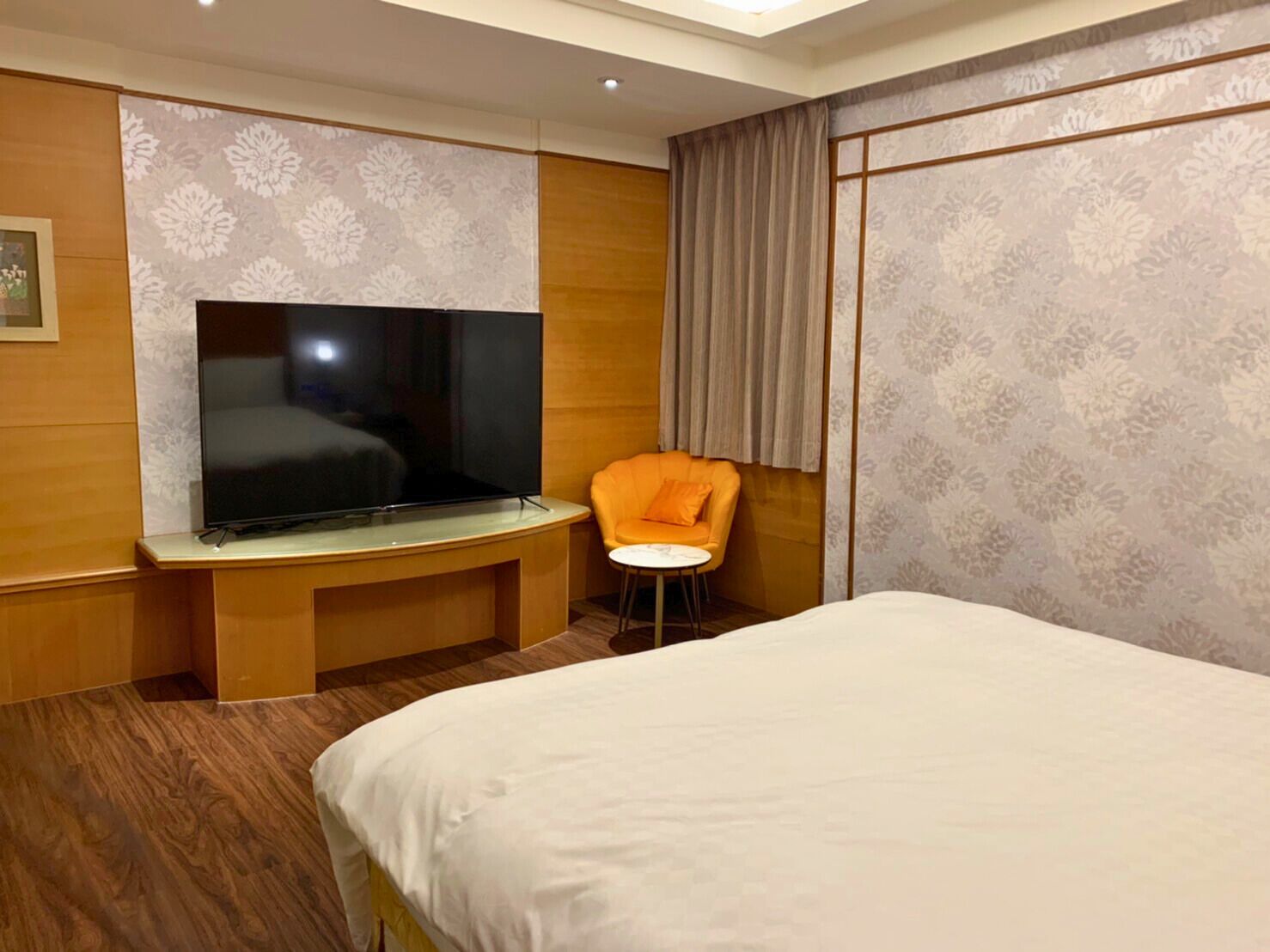 A hotel room with a large bed, a television on a wooden console, an orange chair, and patterned wallpaper.