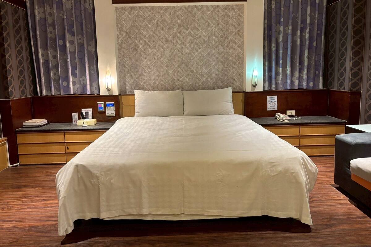 Habitación de hotel con una cama grande, cabecera de madera, mesitas de noche, teléfono y una lámpara de pared.