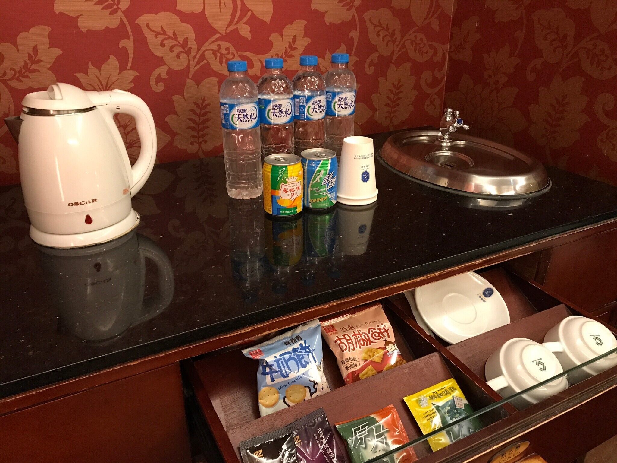A kitchen counter with a white electric kettle, several bottles of water, a can of soda, and a white mug. Below the counter, there is a drawer containing various packaged snacks and a set of white cups.