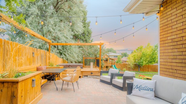 Een buitenpatio met een houten pergola, lichtjes, een eettafel met stoelen en een bank met een kussen waarop 