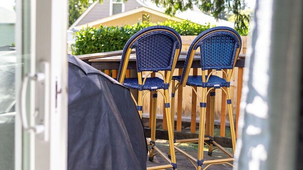 A covered patio area with three blue wicker chairs and a table, surrounded by a wooden fence and greenery.