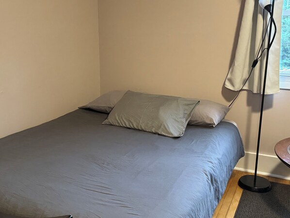 Una cama con ropa de cama gris y dos almohadas, una lámpara de pie y una ventana con cortinas.