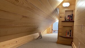 Una habitación estrecha en el ático con listones de madera en el techo y una estantería llena de libros.
