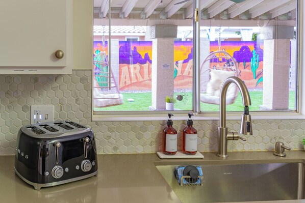 A kitchen with a toaster, two bottles of dish soap, a faucet, and a window with a colorful mural.