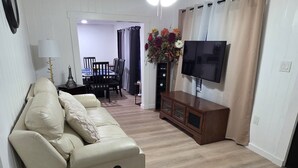 Ein Wohnzimmer mit beiger Couch, einem Holzschrank, einem Flachbildfernseher, einem Esstisch mit Stühlen und einer Blumenkomposition.
