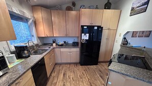 Una cocina con armarios de madera, un refrigerador negro, encimera de granito y ventana con vista a árboles.
