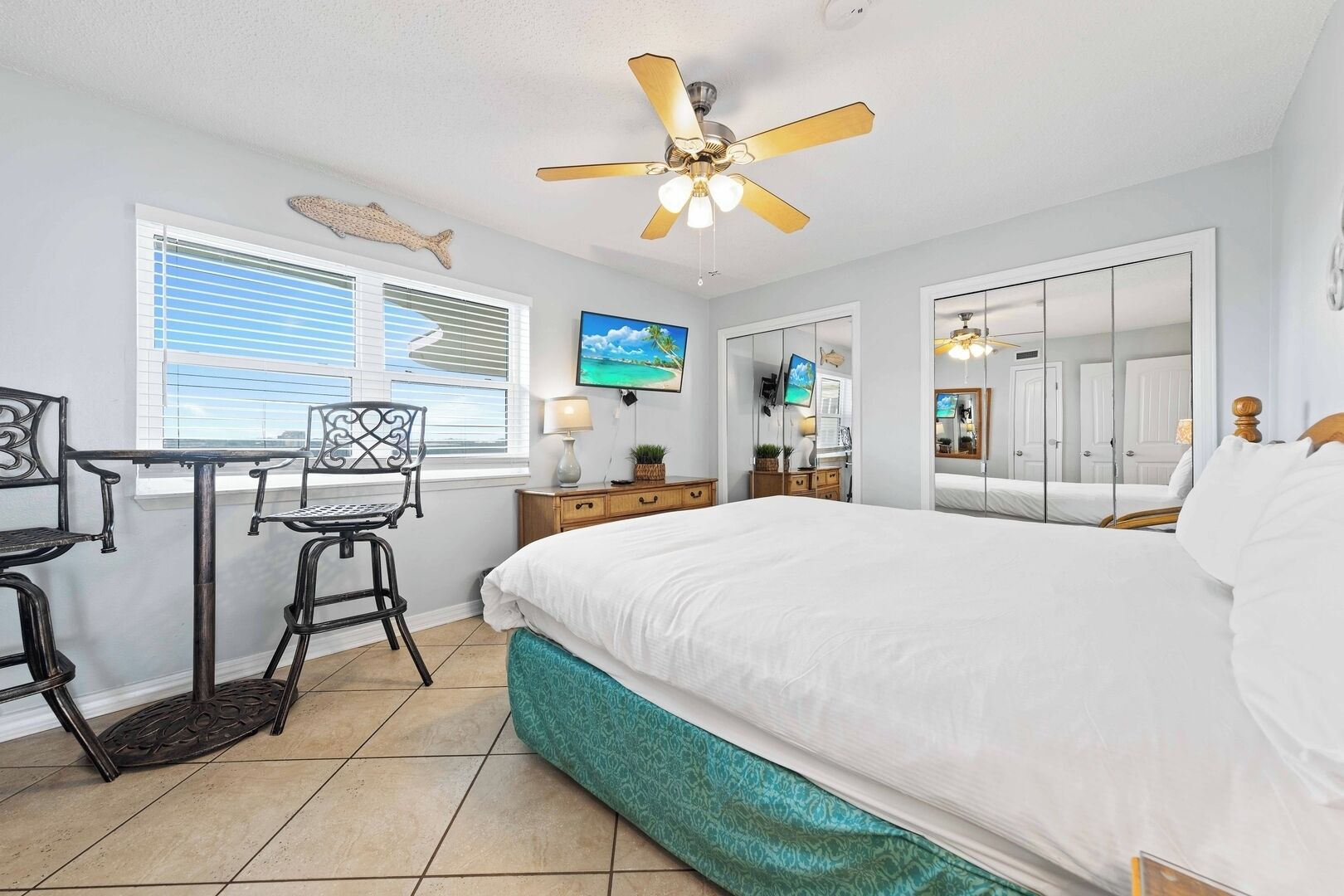 A bedroom with a bed, a desk with two chairs, a ceiling fan, a TV, and a window with blinds.