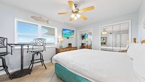 A bedroom with a bed, a desk with two chairs, a ceiling fan, a TV, and a window with blinds.