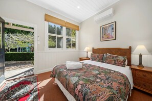 A bedroom with a large bed, two bedside tables with lamps, a window with a view of greenery, and a door leading outside. - Coolangatta (House and Studio) (Katoomba)