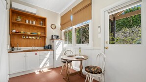 A compact kitchen with white cabinetry, a wooden shelf with dishes, a dining table with chairs, and a window with blinds. - Coolangatta (House and Studio) (Katoomba)
