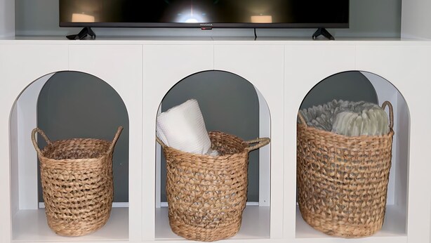 A flat-screen TV mounted on a wall above three woven baskets in a white cabinet.