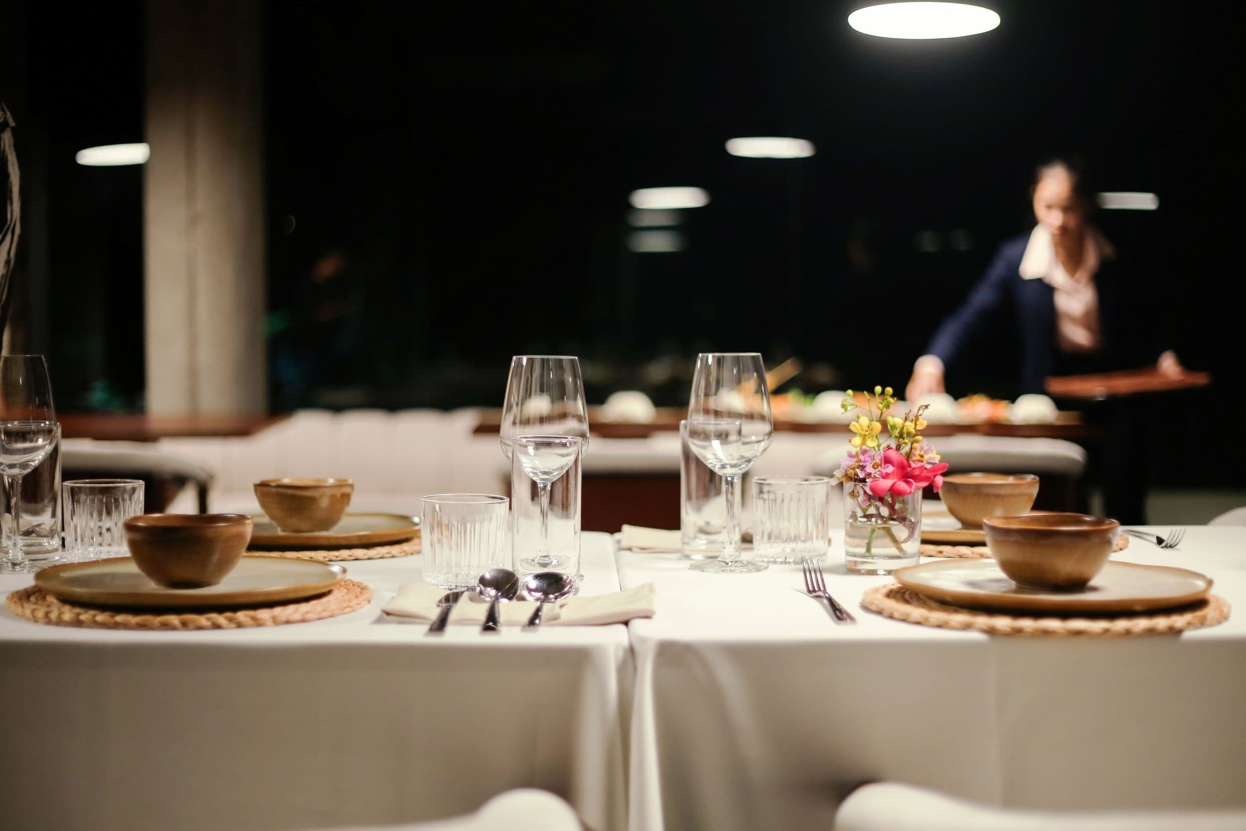 Una mesa dispuesta para comer, con cristalería transparente, platos y un jarrón con flores.