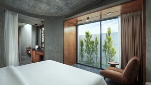 Une chambre d’hôtel moderne avec un grand lit, un bureau en bois, une chaise et une vue sur les arbres par une grande fenêtre.