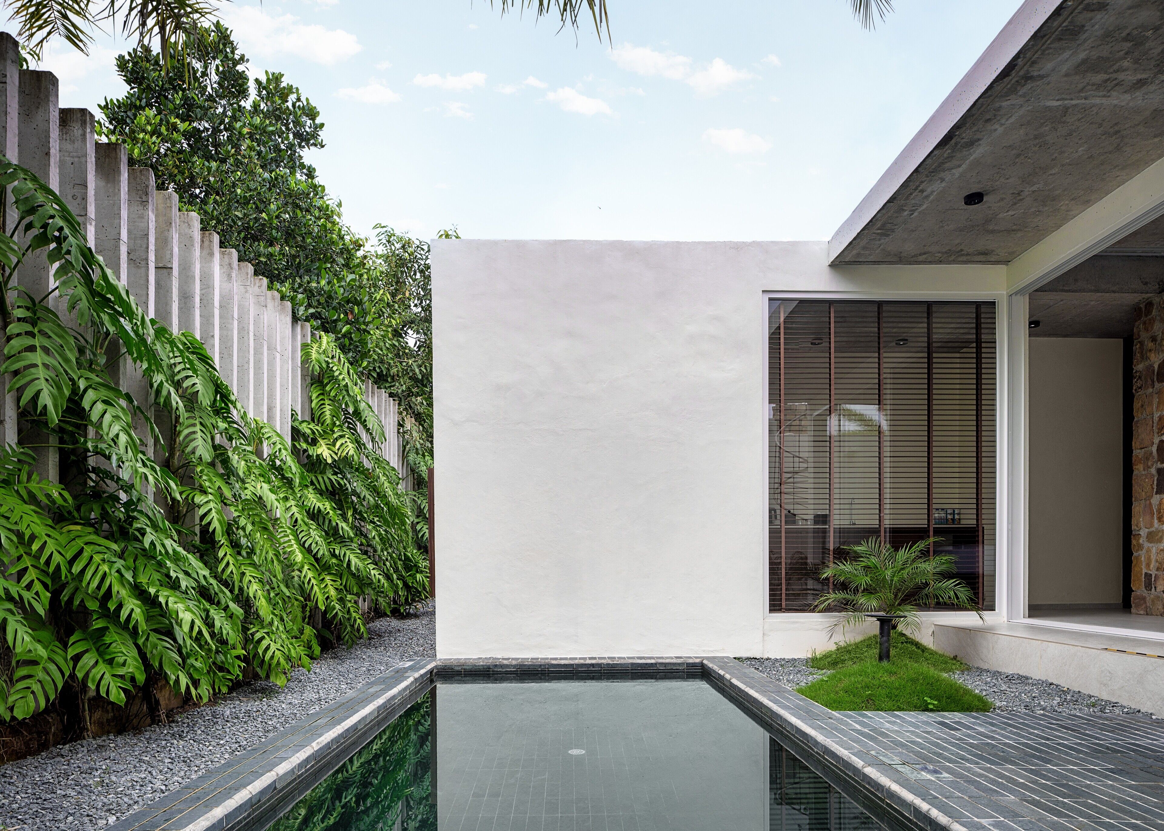 Un área de piscina exterior moderna con un muro de concreto, un pequeño jardín y un edificio con un gran ventanal.