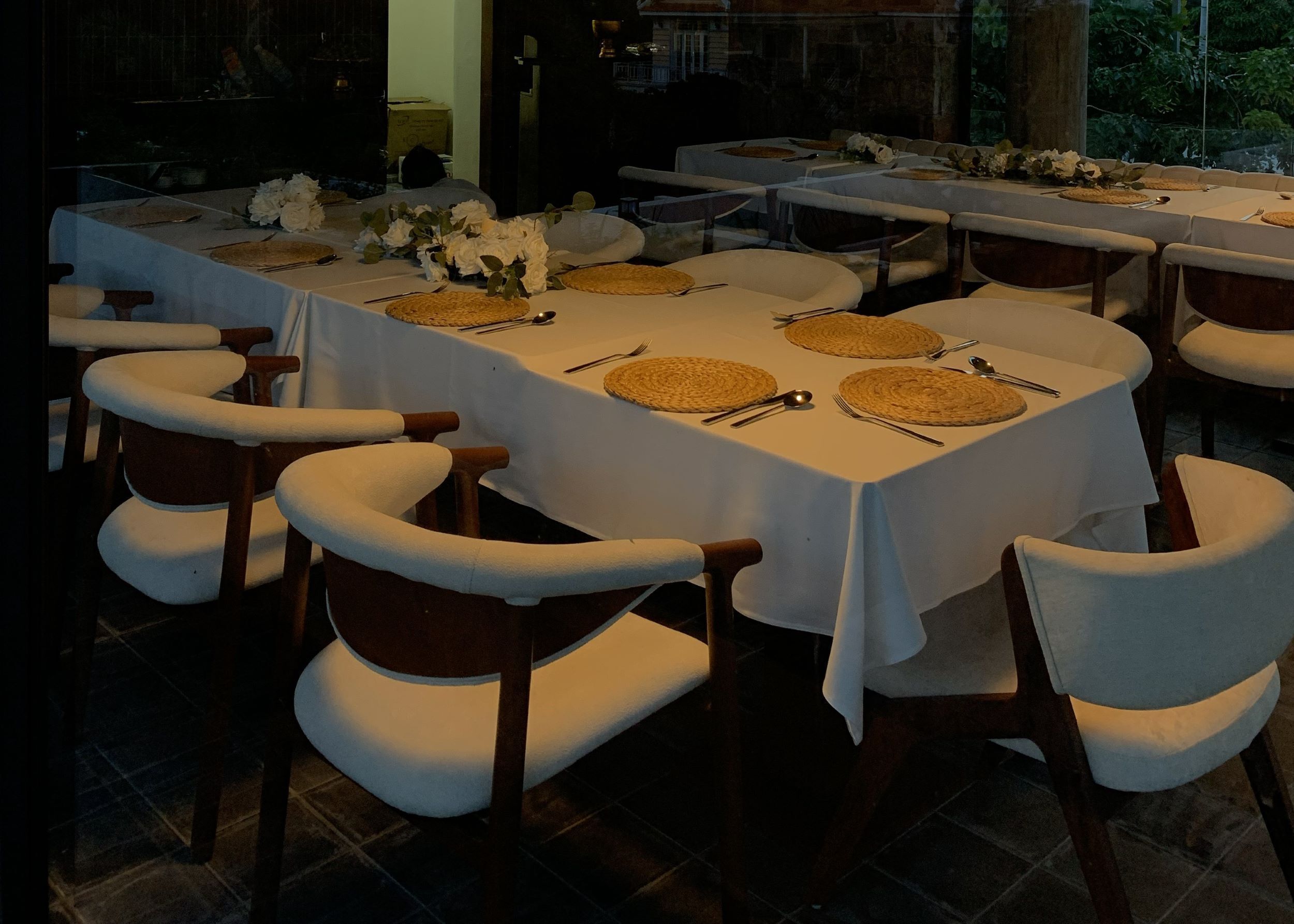 A dining area with multiple tables set with white tablecloths, woven placemats, and white chairs.