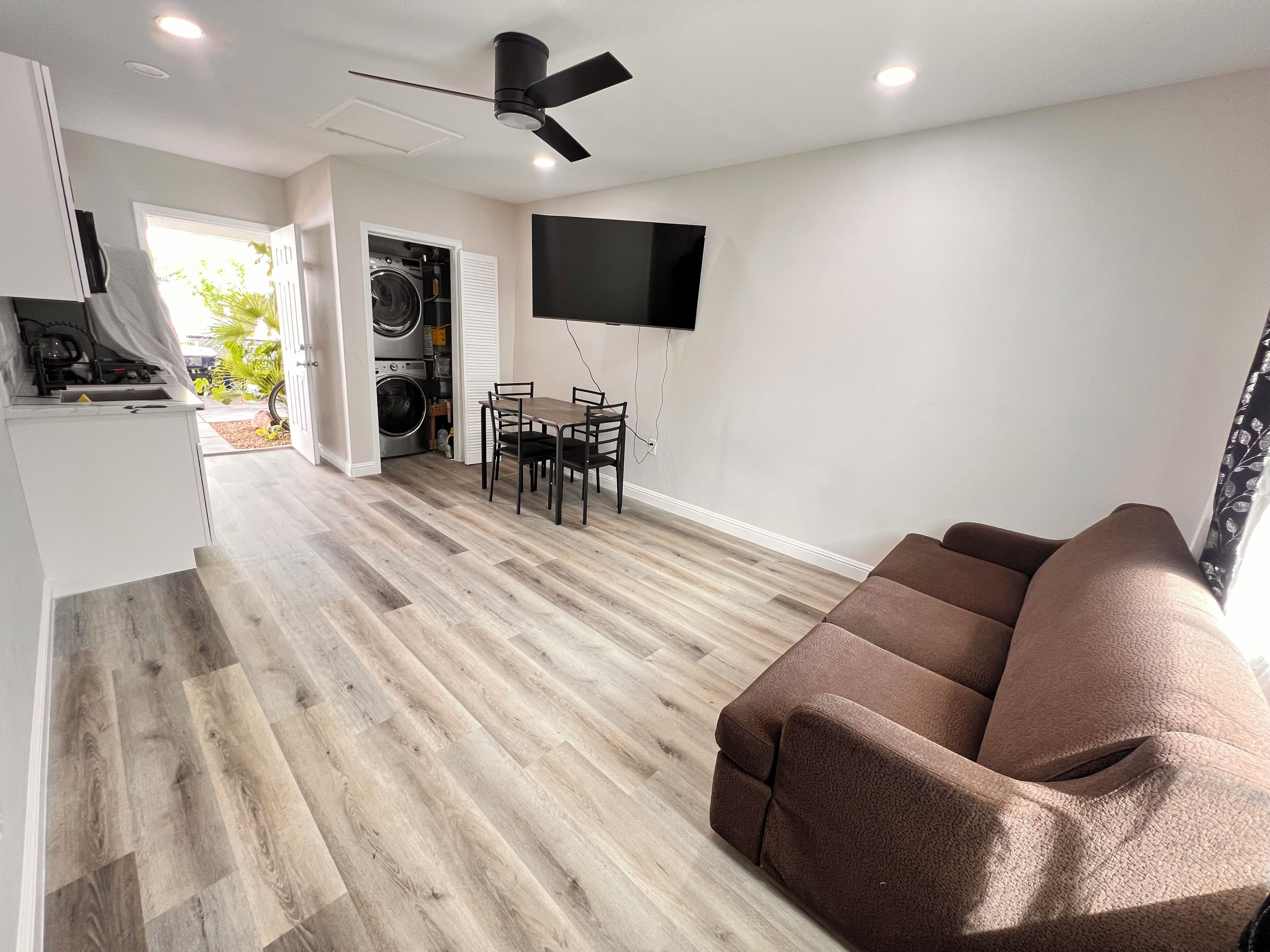 A modern living room with a brown sofa, a dining table with chairs, a ceiling fan, a flat-screen TV mounted on the wall, and a laundry area with a washing machine and dryer.