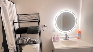 A bathroom with a toilet, a sink, a mirror, and a shelf with towels. - Tranquil Beautiful Studio (Las Vegas)