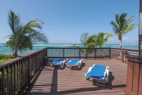 A wooden deck with three sun loungers facing the ocean, surrounded by palm trees and a clear view of the sea.