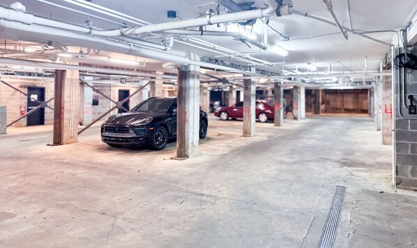 Un estacionamiento con pisos y columnas de concreto, un coche negro estacionado al frente y un coche rojo al fondo.