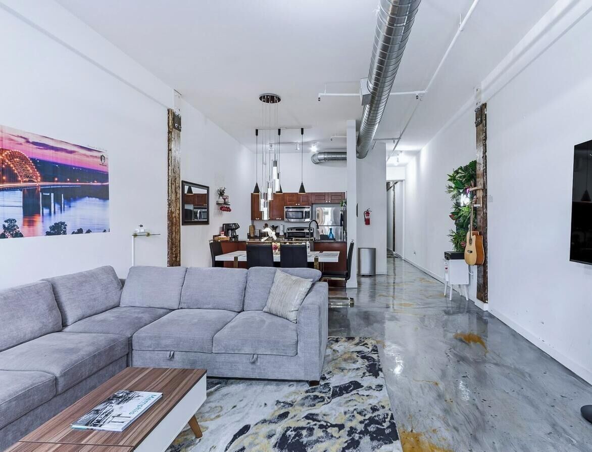 A modern living room with a grey sofa, a coffee table, and a dining area with wooden cabinets and a kitchen in the background.