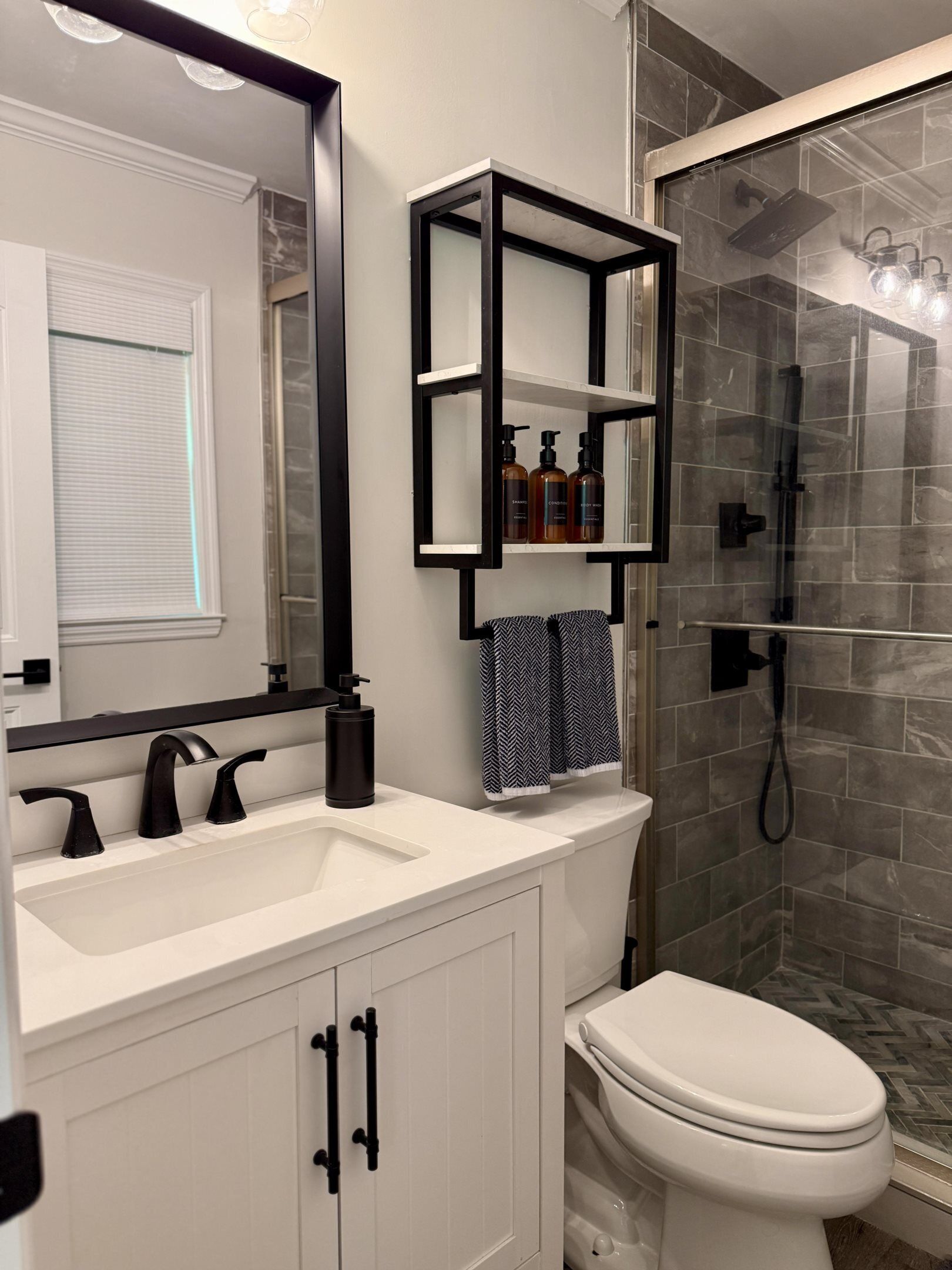 Ein modernes Badezimmer mit einem weißen Waschbecken, schwarzen Wasserhähnen, einer weißen Toilette, einer Duschkabine aus Glas und einem schwarzen Regal mit Pflegeprodukten.