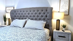 Un lit bien fait, recouvert d’une couvre-lit à motifs, avec une tête de lit capitonnée, deux tables de chevet dotées de lampes et, au mur, un cadre contenant une citation inspirante.