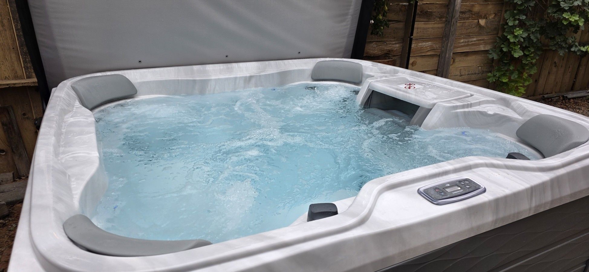 A hot tub with a control panel, surrounded by a wooden fence and foliage.