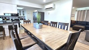Une cuisine moderne dotée d’un îlot central, un coin repas avec une longue table et des chaises, et un salon avec un canapé et un pouf.