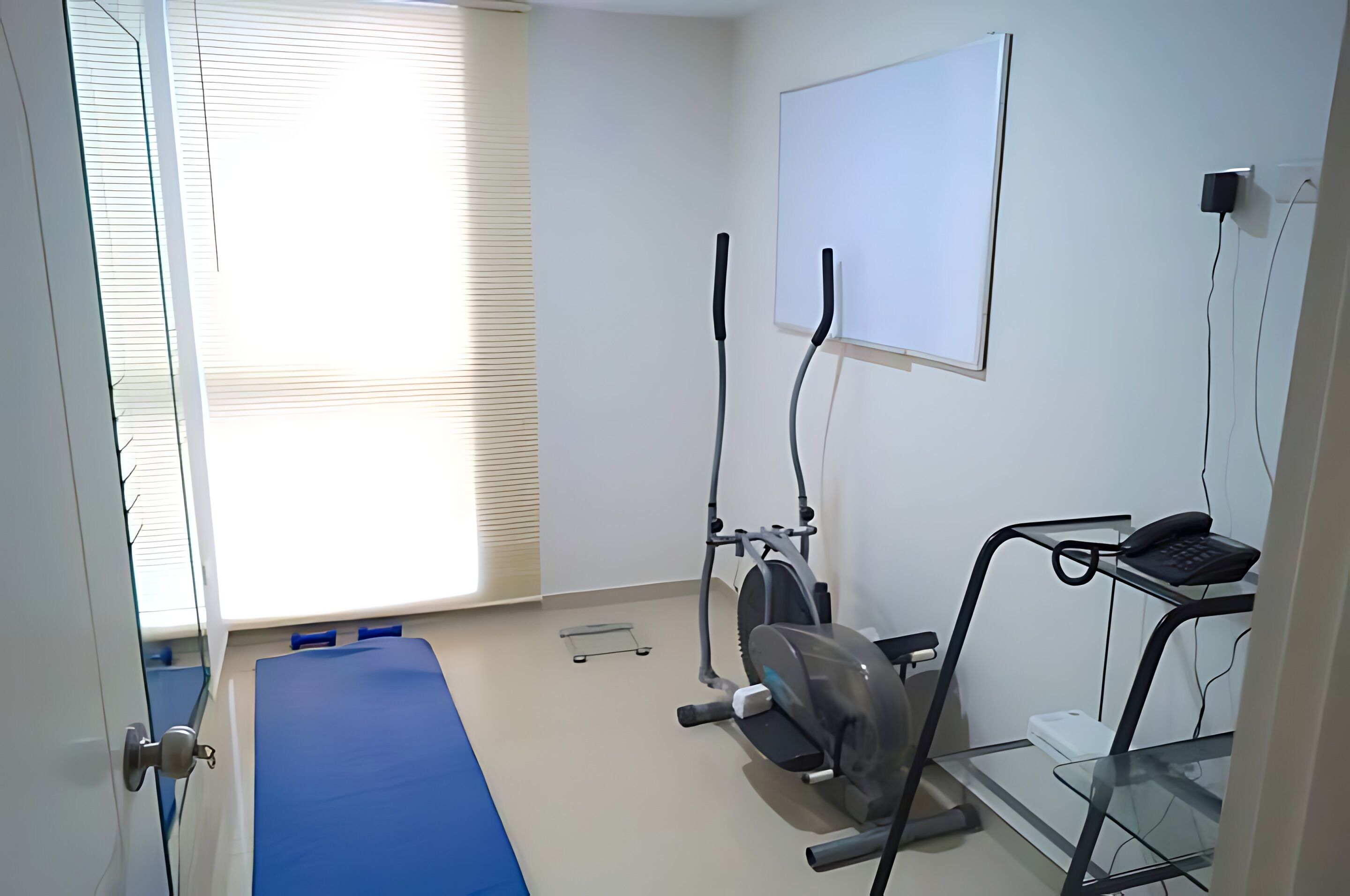 A room with a treadmill, a blue mat, a whiteboard, and a desk with a phone.
