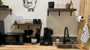 Une cuisine compacte équipée d’un four à micro-ondes, d’une machine à café et d’un évier. Des étagères en bois, situées au-dessus du plan de travail, servent à ranger des tasses et présentent un coq décoratif.