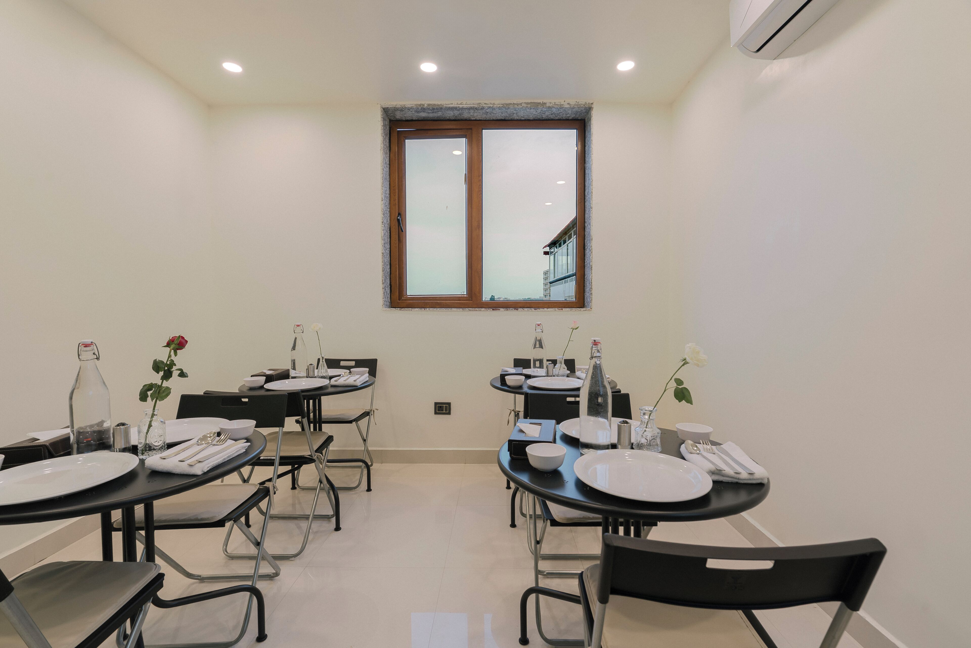 A dining area with tables set for four, each with plates, cutlery, and a bottle. There are chairs around the tables, and a window above the tables.