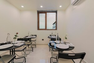 A dining area with tables set for four, each with plates, cutlery, and a bottle. There are chairs around the tables, and a window above the tables.