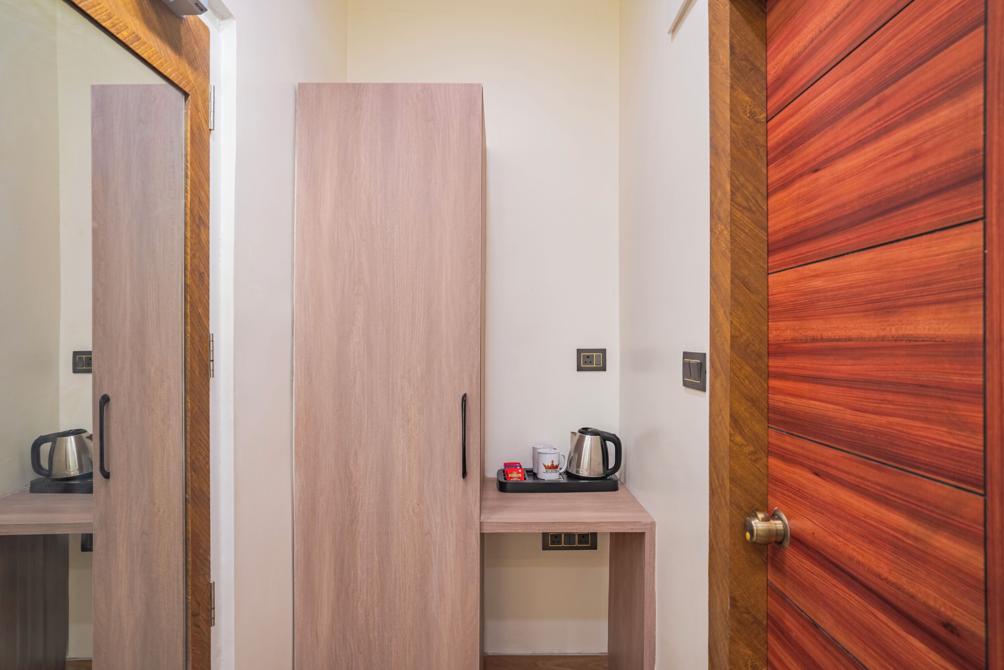 A room with a wooden wardrobe, a small table with a kettle and a mug, and a door with a wooden panel.