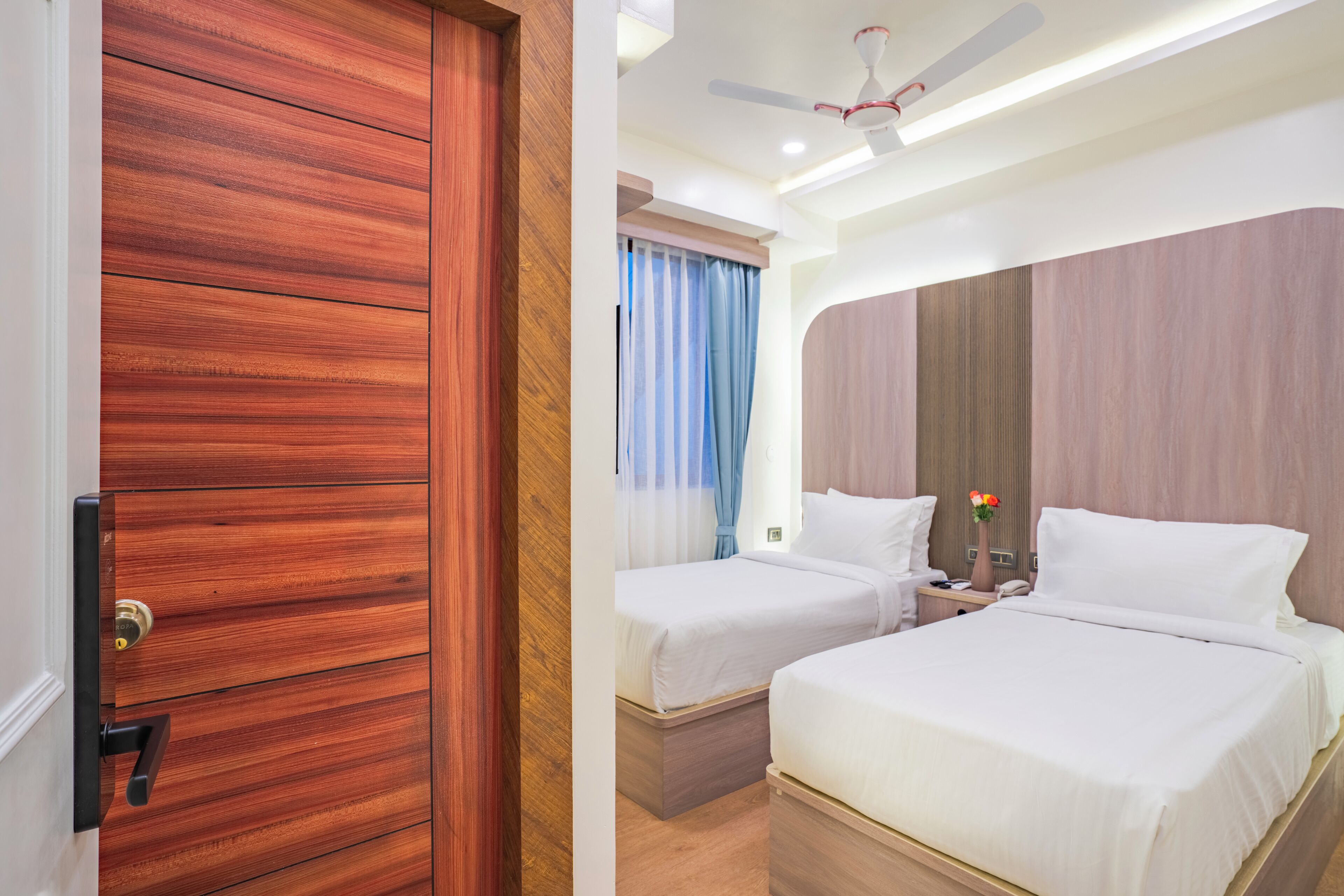 A hotel room with two beds, a wooden door, a ceiling fan, and a nightstand with a vase of flowers.