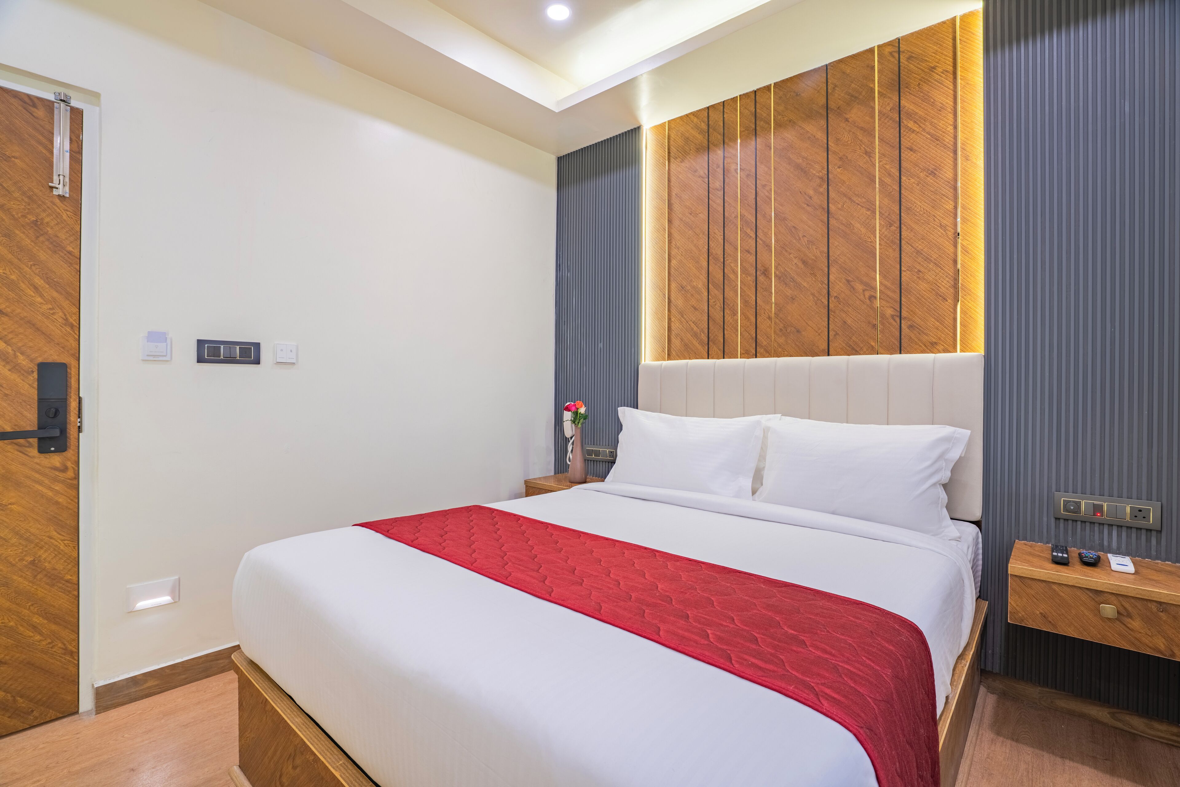 A modern hotel room with a large bed, a wooden headboard, a nightstand, and a wall-mounted control panel.