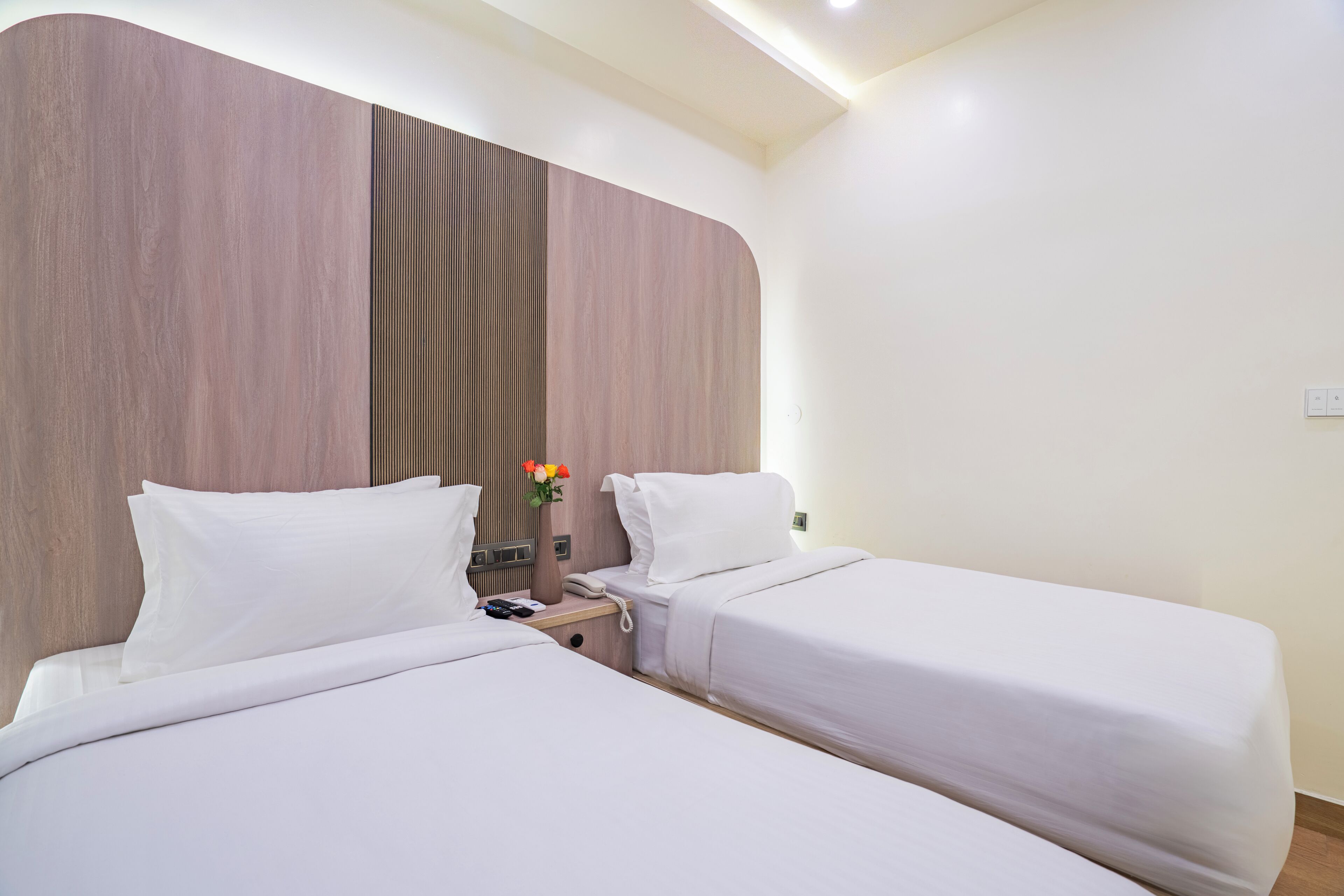 Een hotelkamer met twee enkele bedden, een houten hoofdbord, een nachtkastje met een telefoon en een vaas bloemen, en een witte muur.