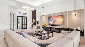 A modern living room with a sectional sofa, a coffee table, a TV mounted on the wall, and a fireplace. - Bluewater- 4 bedroom, 4 bath luxury Villa - Vesteva (Cape Coral)