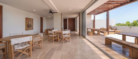 A spacious indoor area with wooden tables and benches, large windows, and a ceiling fan.