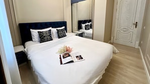 Un lit bien fait, avec une couette blanche et des coussins décoratifs, une table de chevet sur laquelle se trouvent un livre et des fleurs, et un grand miroir au-dessus du lit.