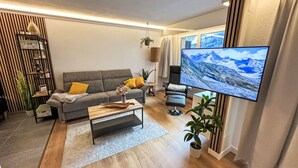 En moderne stue med en grå sofa, et træsofabord og et stort fladskærms-tv, der viser et bjerglandskab.