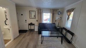 Una sala de estar con un sofá negro, una mesa de centro de vidrio, una mesita auxiliar con una lámpara, un radiador y una ventana con cortinas.