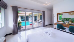A hotel room with a large bed, a desk with two chairs, a large window with curtains, and a view of a swimming pool. - Natcha villa 41 (Pattaya)