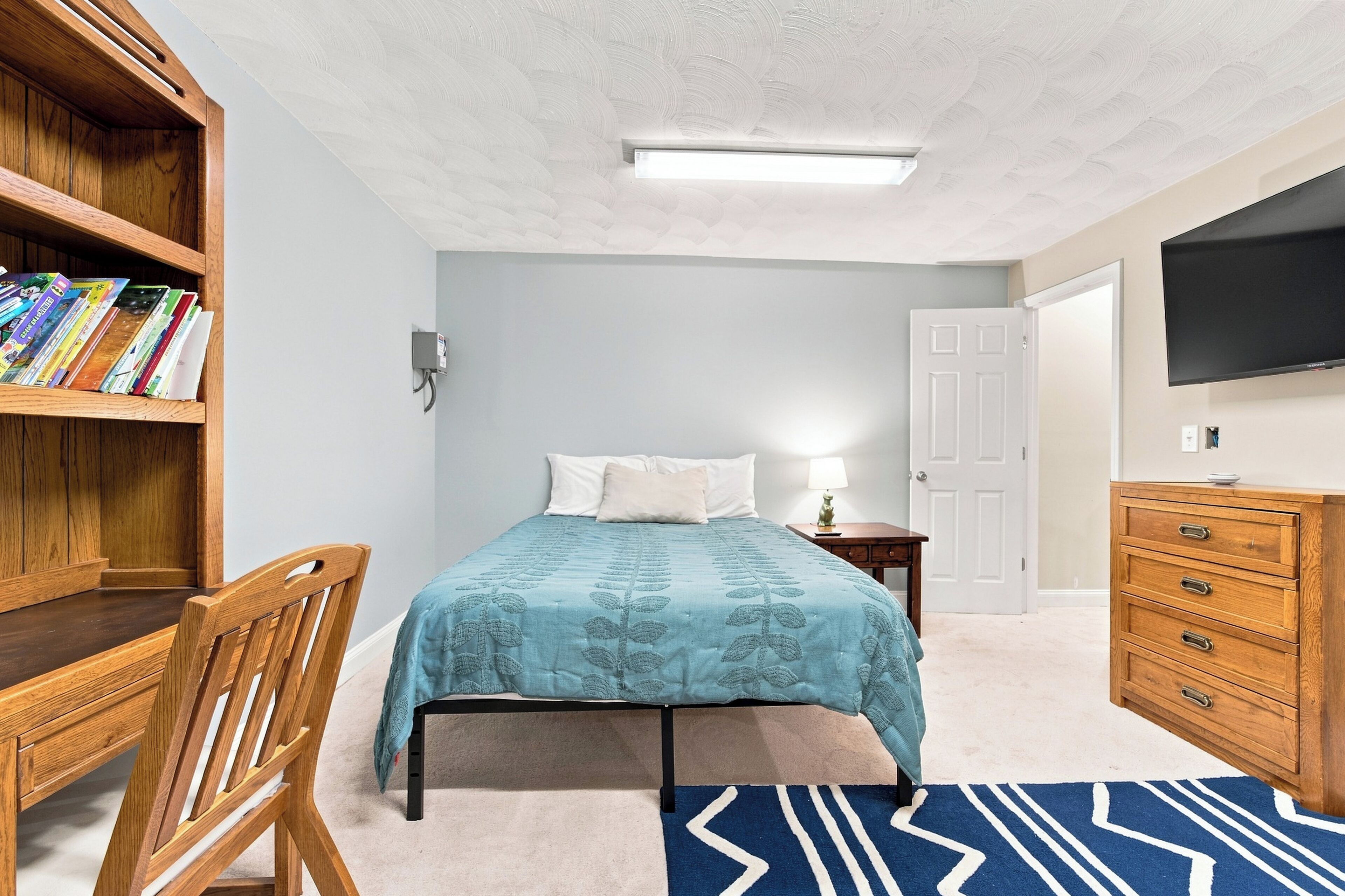 A bedroom with a bed, a wooden bookshelf, a desk, a chair, a dresser, a television, and a patterned rug.