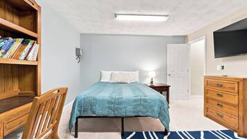 A bedroom with a bed, a wooden bookshelf, a desk, a chair, a dresser, a television, and a patterned rug.