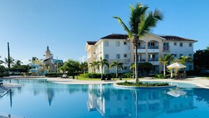 A large swimming pool with a clear blue water surface, surrounded by a sandy area and palm trees. In the background, there is a multi-story building with balconies and a water slide structure. - Studio with beach and pool access at Club Cadaqués, Bayahibe (San Rafael de Yuma)