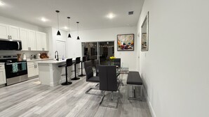 Una cocina moderna con armarios blancos, encimera negra y una isla central con taburetes. Hay un comedor con una mesa de vidrio y sillas, y un cuadro grande en la pared.