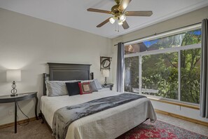 Une chambre à coucher avec un lit, une table de chevet, un ventilateur de plafond, une fenêtre donnant sur des arbres et un tapis rouge.