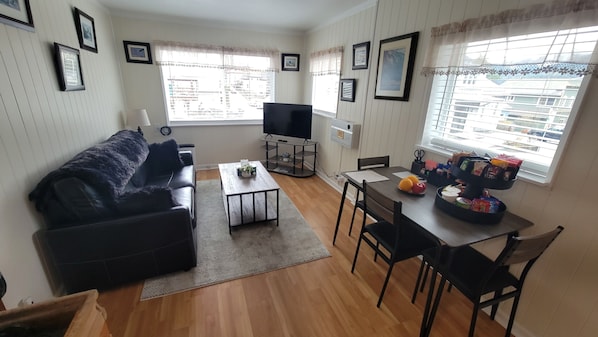 A living room with a black sofa, a TV on a stand, a dining table with chairs, and a window with blinds.