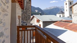 Ein hölzerner Balkon mit Steinmauer und Metallrinne, mit Blick auf eine gebirgige Landschaft mit einem Kirchturm.
